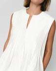 A woman wears the SUNCOO Conely Dress, a sleeveless white pleated summer dress with a relaxed fit and split neckline. She pairs it with shoulder-length brown hair and black sunglasses against a plain, light background.