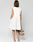 A woman with shoulder-length hair faces away, wearing the SUNCOO Conely Dress—a relaxed fit, sleeveless white summer dress—and white heels, holding a beige and black striped tote bag against a plain light background.