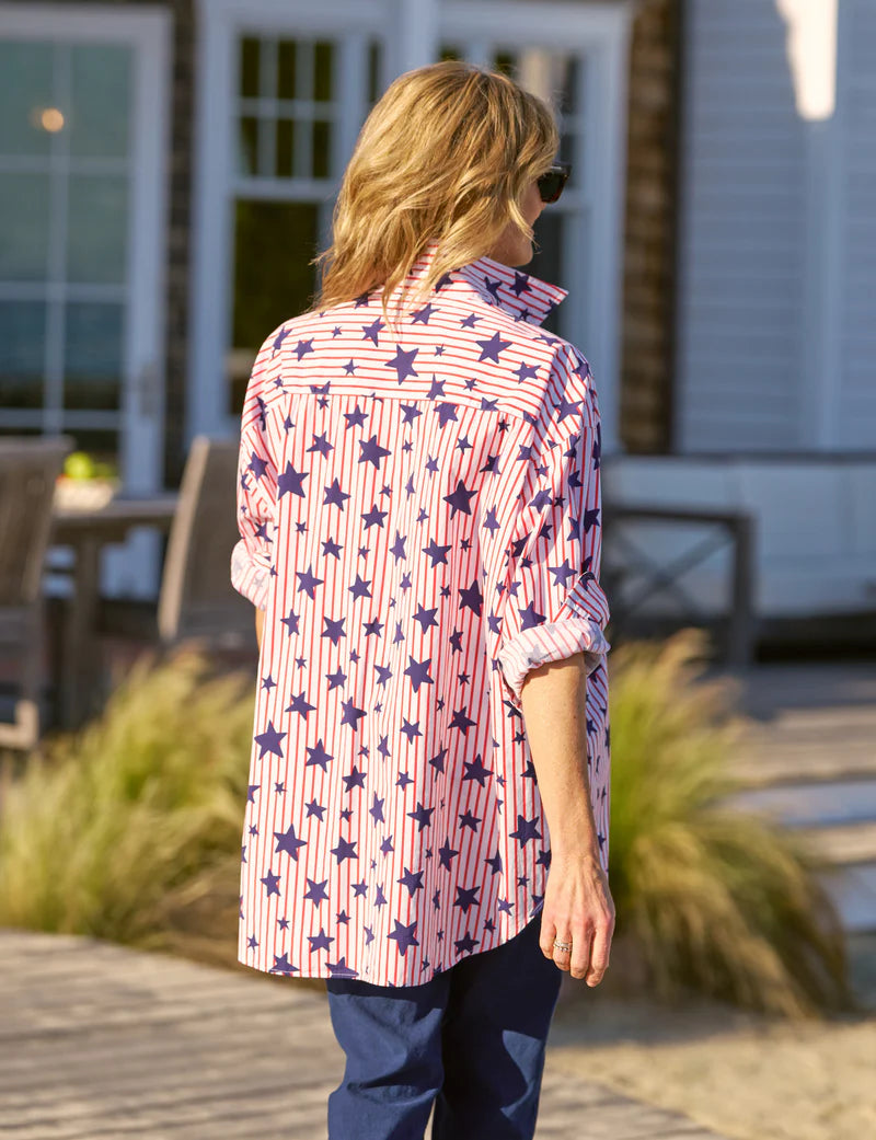 A person with blonde hair, wearing sunglasses and blue pants, models the Frank & Eileen Eileen S25 relaxed fit long-sleeved shirt with a pink and purple star pattern outdoors near a patio and greenery.