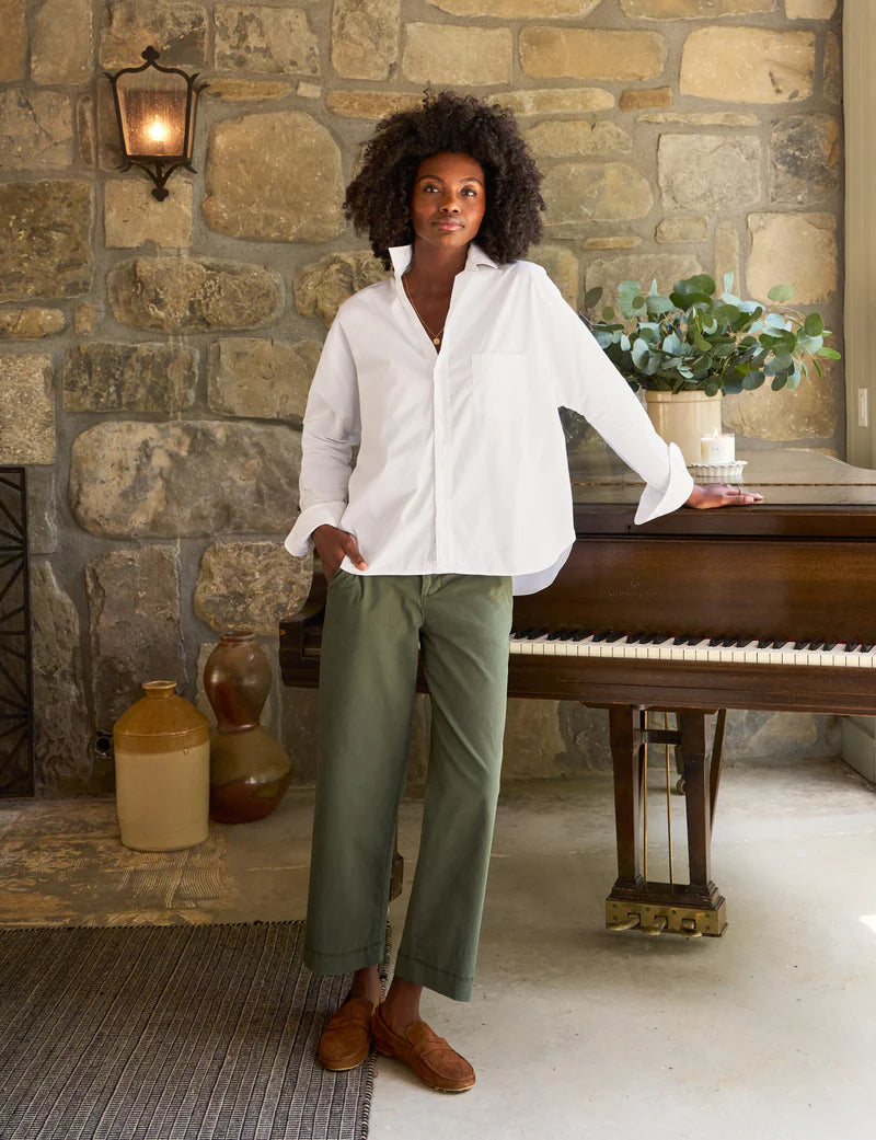 A person with curly hair, wearing a relaxed-fit, 100% cotton Oona Continuous Button-Up Shirt by Frank & Eileen and green pants, stands with one hand resting on a piano in a room with stone walls. There is a plant on the piano and two large vases on the floor nearby. A lit wall sconce adds warm light to the scene.