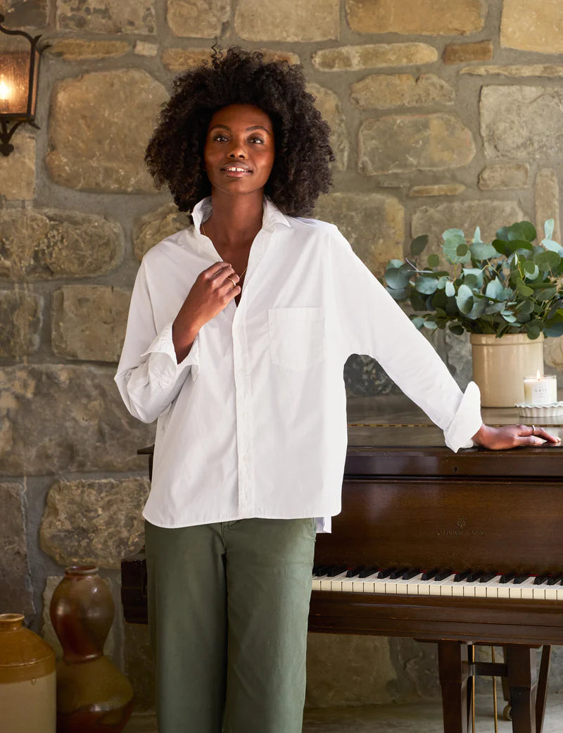 A person stands indoors, leaning against a piano in front of a stone wall. They wear the Oona Continuous Button-Up Shirt from Frank & Eileen, which is a relaxed fit, 100% cotton white shirt, along with green pants. Their curly hair is loose, and they smile slightly. The top of the piano holds a vase with green foliage and a few small decorative items.