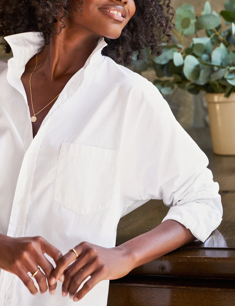 A person with curly hair is leaning against a wooden surface. They are wearing the Oona Continuous Button-Up Shirt made from 100% cotton in white, by Frank & Eileen, with the collar popped up and sleeves rolled. The person also wears gold rings and a delicate gold necklace. A potted plant is blurred in the background.