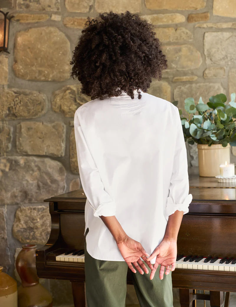 A person with curly hair is standing with their back turned, facing a stone wall and an upright piano adorned with decorative plants on top. They are wearing a Frank & Eileen Oona Continuous Button-Up Shirt made from 100% cotton and green pants, with their hands clasped behind their back.