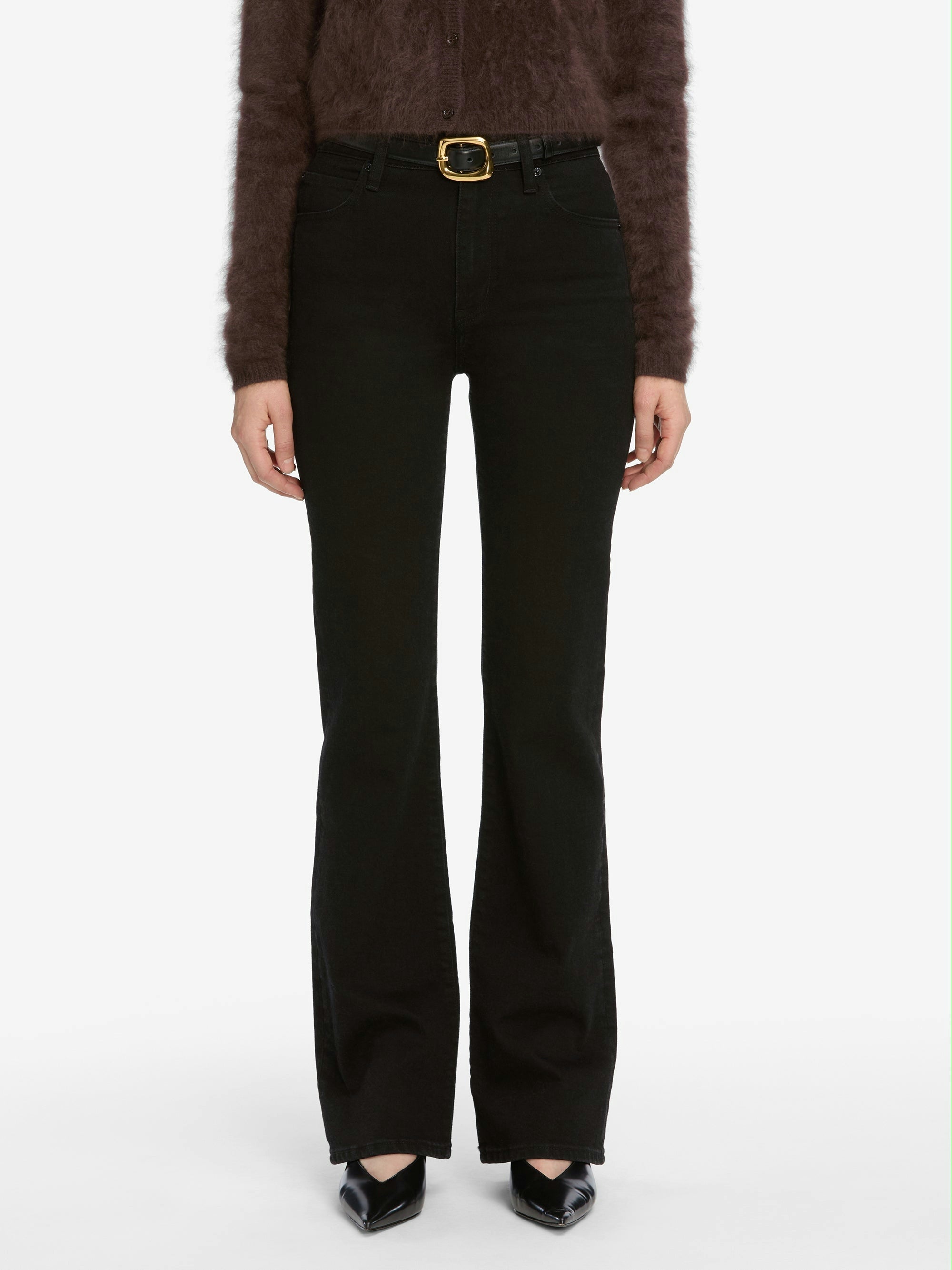 A person wears black bootcut jeans, a fuzzy dark sweater, and pointed black heels, styled with FRAME's The Organic Buckle Belt featuring a gold buckle, all set against a white background.