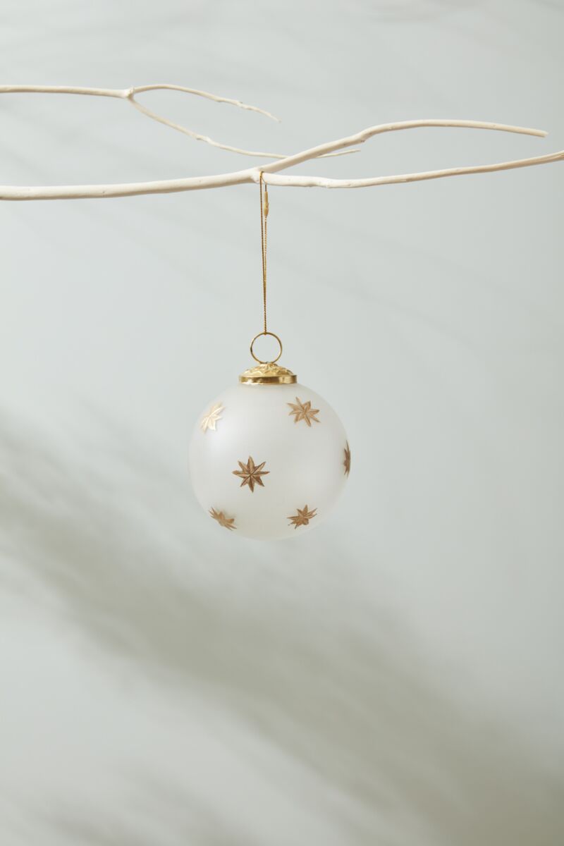The Accent Decor North Star Ornament 4", featuring gold star designs on white, hangs from a bare branch against a neutral background. Its 4-inch size fits perfectly in a 4x4x4 inch box.