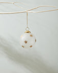 The Accent Decor North Star Ornament 4", featuring gold star designs on white, hangs from a bare branch against a neutral background. Its 4-inch size fits perfectly in a 4x4x4 inch box.