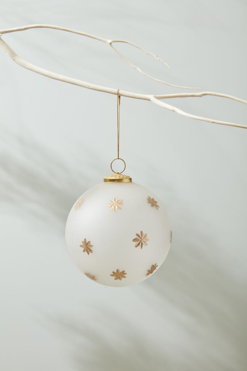 The North Star Ornament 6" by Accent Decor features frosted white glass with gold star patterns, ideal for gifting in a 6x6x6 box, and looks stunning hanging from a white branch against a light background.