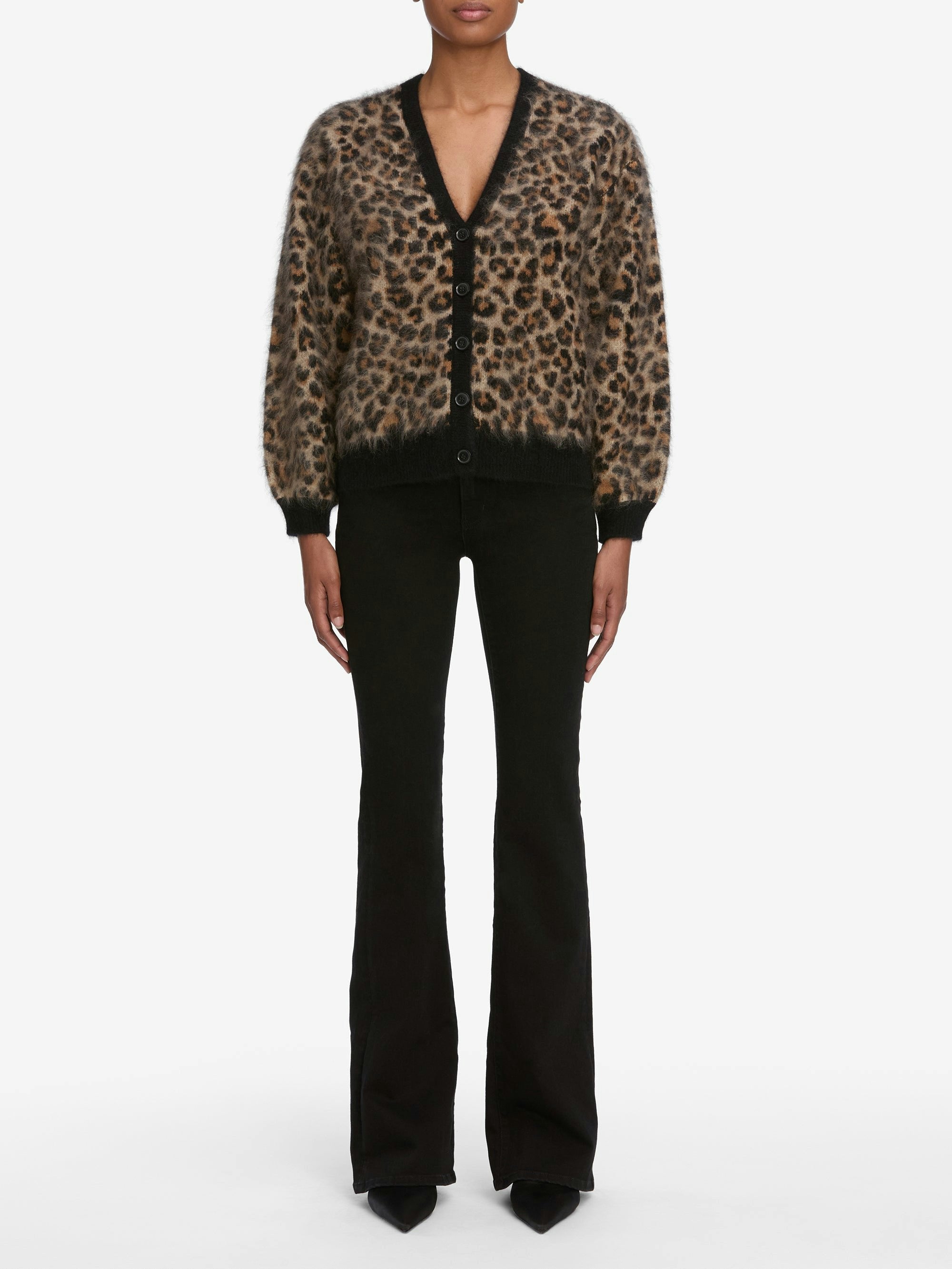 A person wearing the FRAME Brushed Leopard Cardi, featuring bold leopard-print and black trim, paired with black flared pants and pointed-toe shoes, stands against a plain white background.