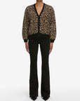 A person wearing the FRAME Brushed Leopard Cardi, featuring bold leopard-print and black trim, paired with black flared pants and pointed-toe shoes, stands against a plain white background.
