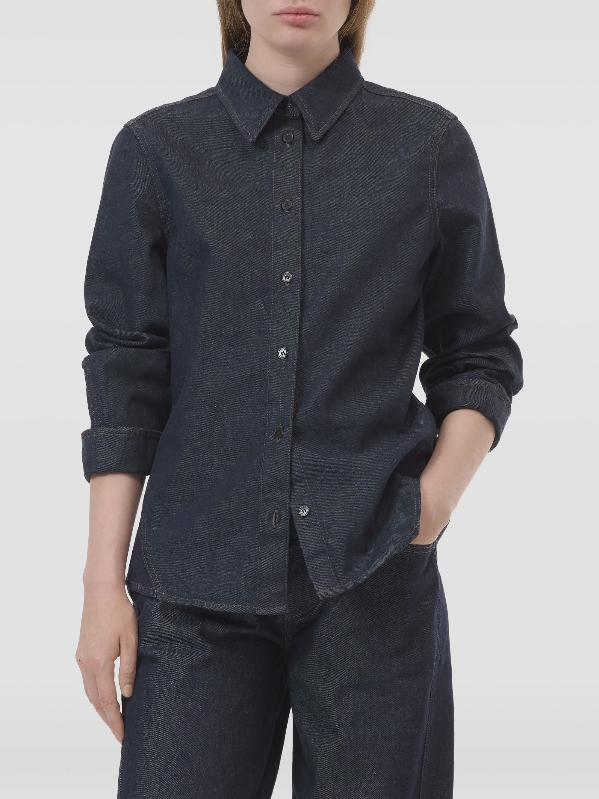 A person wears the CLOSED Seaming Shirt, a dark blue cotton denim long-sleeve button-up, paired with matching pants, standing with one hand in their pocket against a plain background.