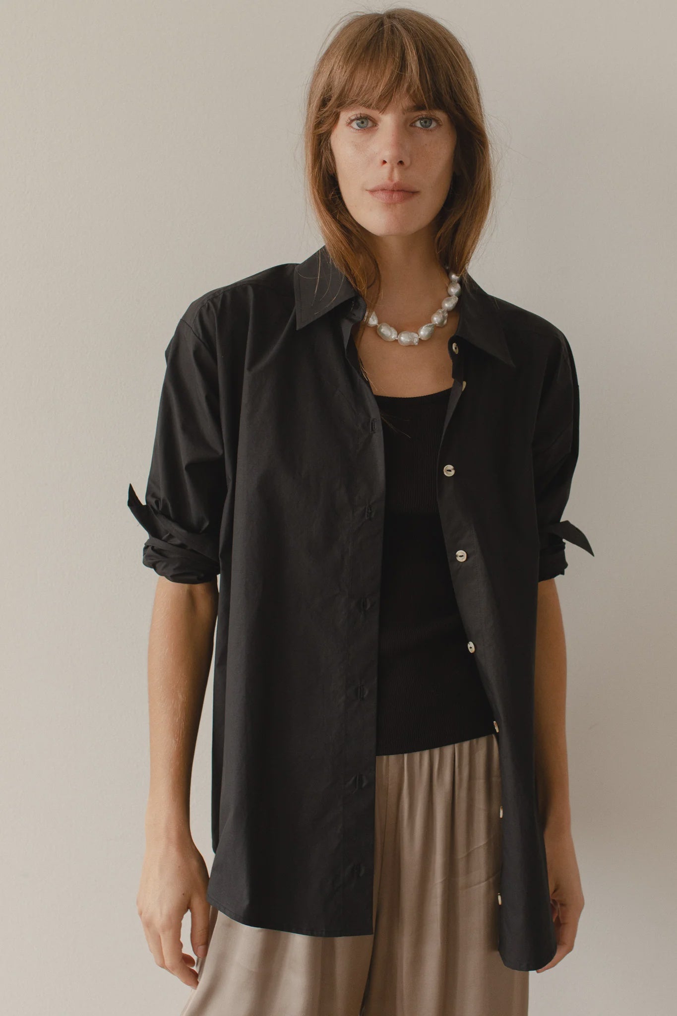 A person with long hair stands against a plain background, wearing Donni's The Pop Shirt—black cotton poplin featuring tortoise-like buttons—with rolled-up sleeves over a black top and light-colored pants. They accessorize with a pearl necklace and maintain a neutral expression.