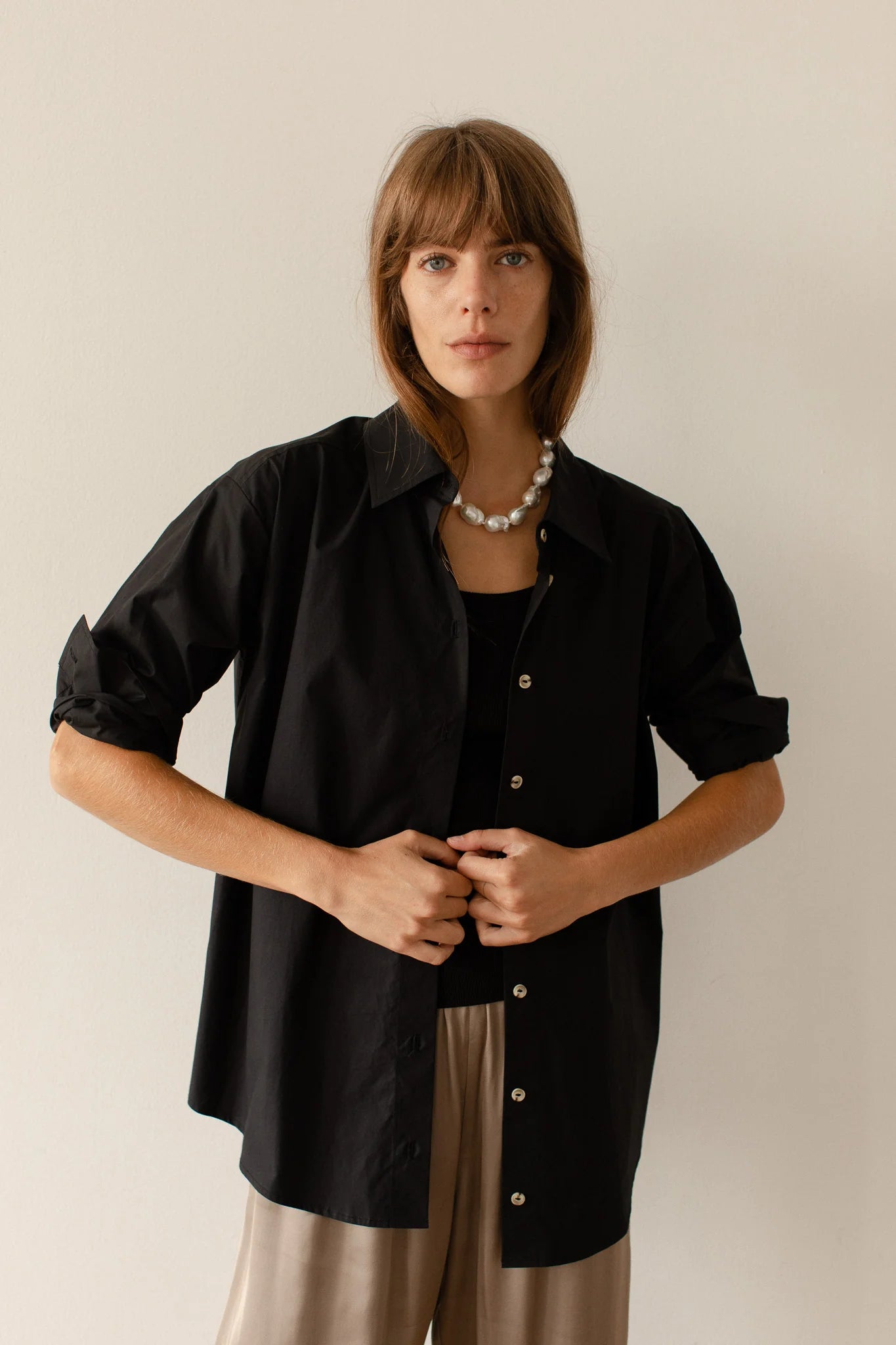 A person with long brown hair stands against a plain background, wearing Donni's The Pop Shirt featuring tortoise-like buttons over a black top, paired with light-colored cotton poplin pants and a pearl necklace. They rest their hands near the shirt's buttons.