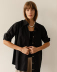 A person with long brown hair stands against a plain background, wearing Donni's The Pop Shirt featuring tortoise-like buttons over a black top, paired with light-colored cotton poplin pants and a pearl necklace. They rest their hands near the shirt's buttons.