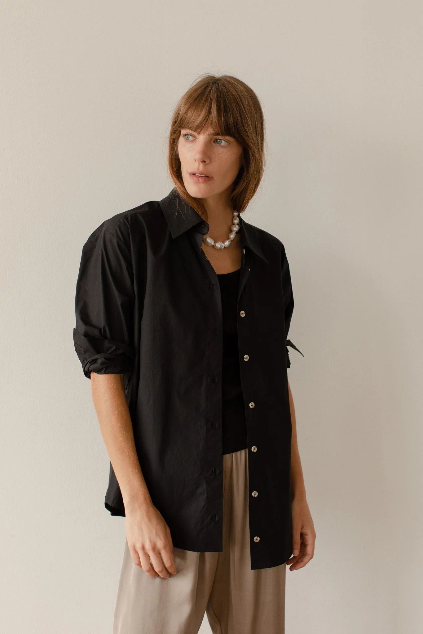 A person with shoulder-length hair wears The Pop Shirt by Donni, a black button-up featuring tortoise-like buttons, over a black top and beige pants. A pearl necklace adorns their neck as they gaze to the side against a plain, light-colored background.