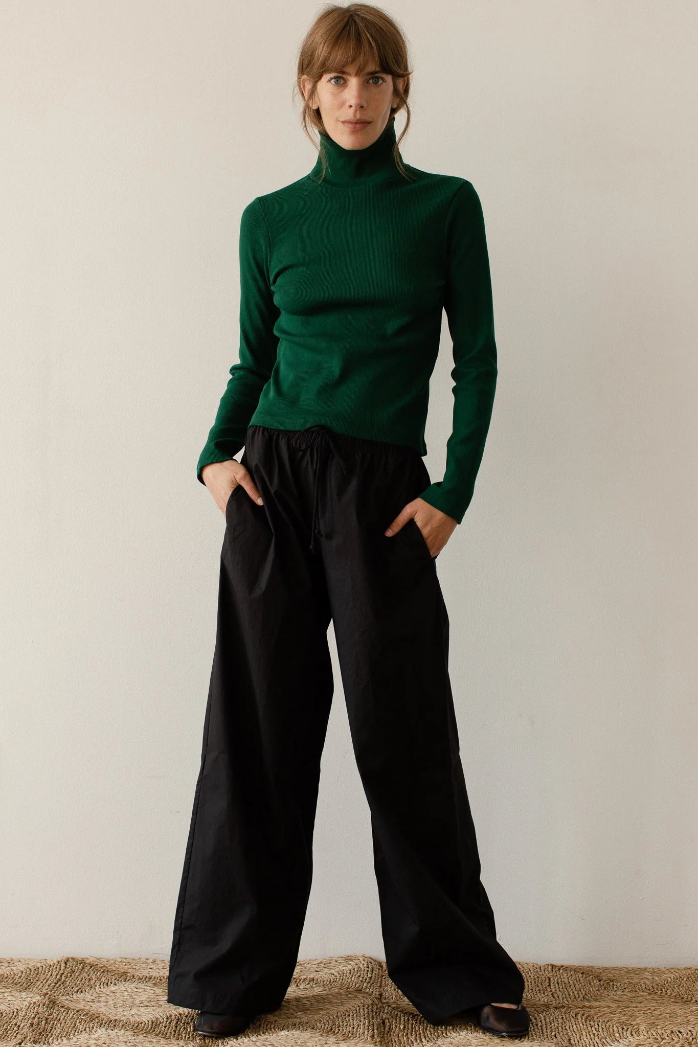 A person stands against a plain background, wearing a green turtleneck sweater and The Pop Pant by Donni in a wide-leg fit. They have their hands in their pockets and are looking at the camera.