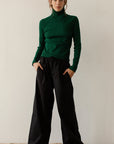 A person stands against a plain background, wearing a green turtleneck sweater and The Pop Pant by Donni in a wide-leg fit. They have their hands in their pockets and are looking at the camera.