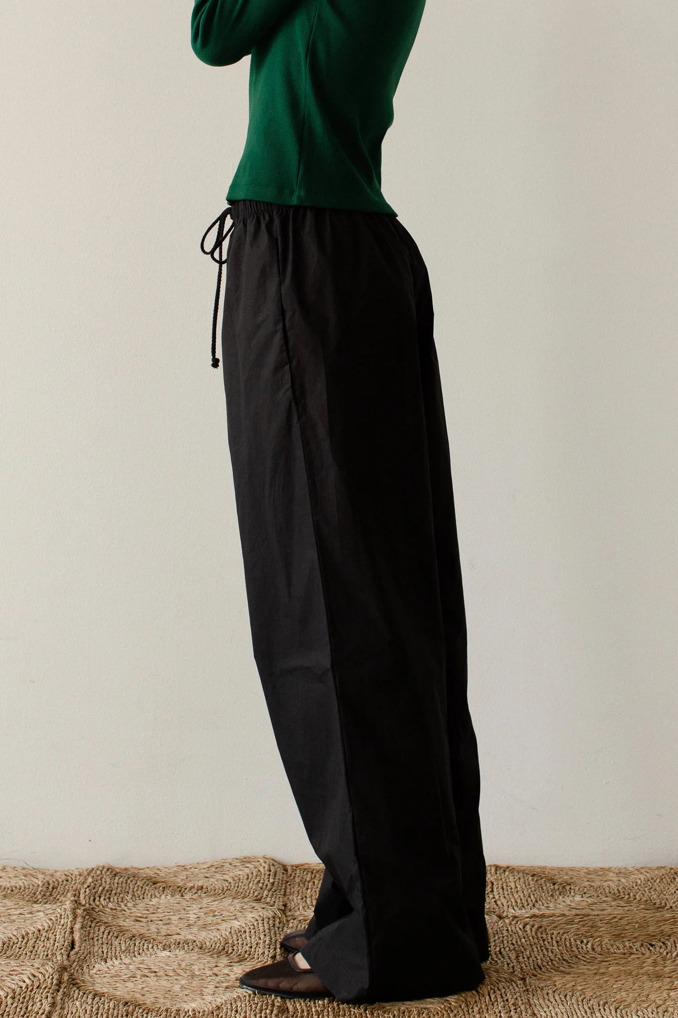 A person stands on a woven rug, wearing a green long-sleeve top and The Pop Pant by Donni in 100% cotton poplin. These wide-leg black pants feature a drawstring waist. The image centers on the outfit, cropping out the person's head and feet.