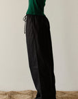 A person stands on a woven rug, wearing a green long-sleeve top and The Pop Pant by Donni in 100% cotton poplin. These wide-leg black pants feature a drawstring waist. The image centers on the outfit, cropping out the person's head and feet.