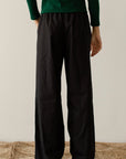 A person stands on a woven rug, wearing a dark green long-sleeve top and The Pop Pant by Donni, made from 100% cotton poplin with a wide-leg fit. Facing away from the camera, their left arm is slightly bent against the backdrop of a plain white wall.