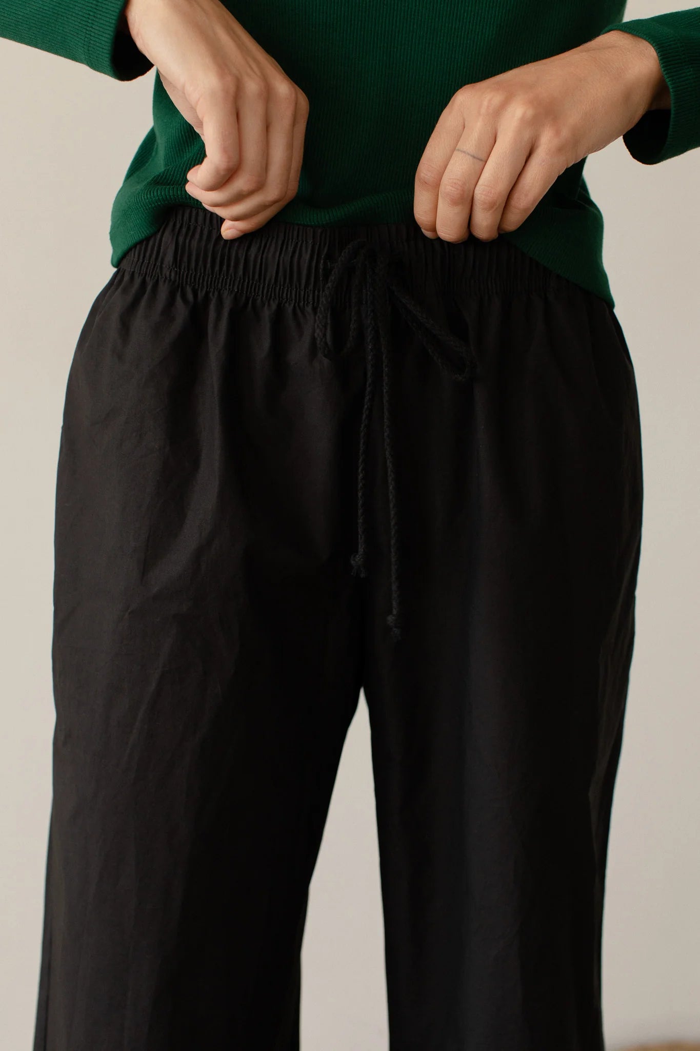 A person in a green top adjusts the drawstring of The Pop Pant, made from 100% cotton poplin by Donni, against a neutral background.