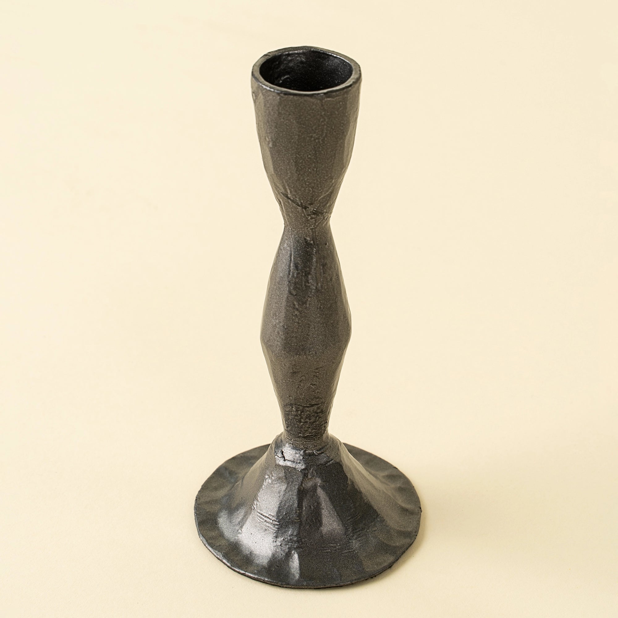 The Zora Forged Candlestick L by Indaba features a dark metallic finish, a textured surface, narrow midsection, and wide flared base, standing upright against a plain light background.