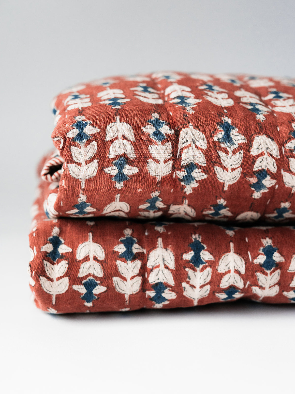 Two folded Blossom Block Print Quilts by Faire, featuring a rust background with a repeating white and blue leaf pattern, stacked on a light surface—perfect for adding bohemian bedding style to your space.