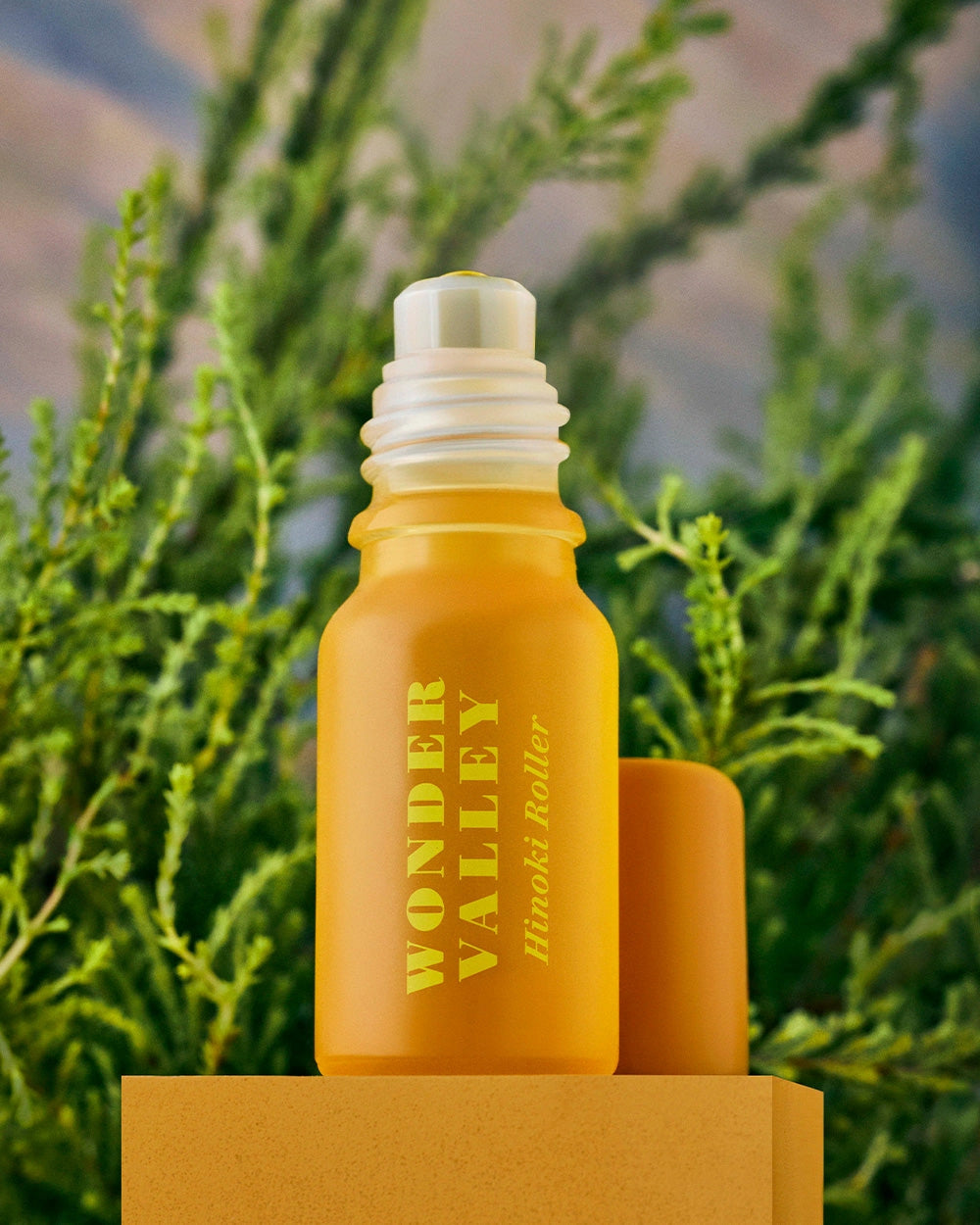 A yellow glass bottle labeled "Faire Hinoki Roll On," an essential oil roller with a hinoki cedar scent, stands on a yellow platform with its cap beside it against a blurred green natural background.