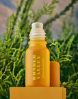 A yellow glass bottle labeled "Faire Hinoki Roll On," an essential oil roller with a hinoki cedar scent, stands on a yellow platform with its cap beside it against a blurred green natural background.