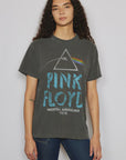 A person with long, curly hair is wearing a dark gray Pink Floyd North American Tour tee by People Of Leisure. The shirt is made from organic cotton and showcases a prism design with a rainbow. They stand against a plain background and pair it with dark jeans.