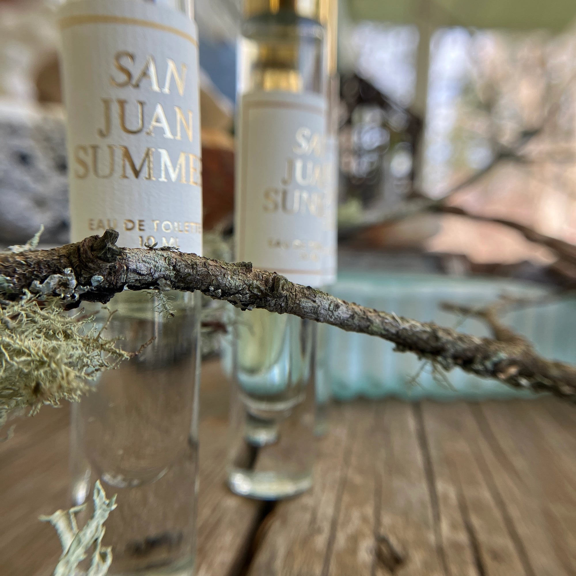 Close-up of two Faire San Juan Roll On perfume bottles on wood, with a lichen-covered twig in front and a blurred backdrop—evoking the cedar and spice scents of the San Juan Islands.