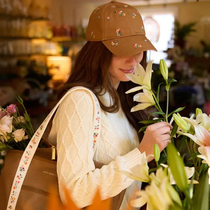 A woman in a cream knit sweater stands in a flower shop, wearing the Faire Ditsy Dahlia Baseball Cap and holding a large tan bag with flowers as she gently smells a bouquet of white lilies among colorful blooms.