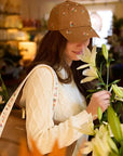 A woman in a cream knit sweater stands in a flower shop, wearing the Faire Ditsy Dahlia Baseball Cap and holding a large tan bag with flowers as she gently smells a bouquet of white lilies among colorful blooms.