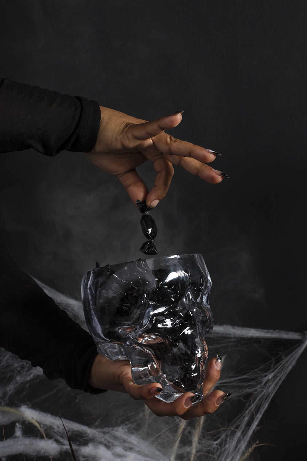 A hand with dark nails drops a black crystal into the Accent Decor Reaper Pot, a skull-shaped glass terrarium shrouded in smoke and cobwebs—ideal for spooky decor on a mysterious dark background.