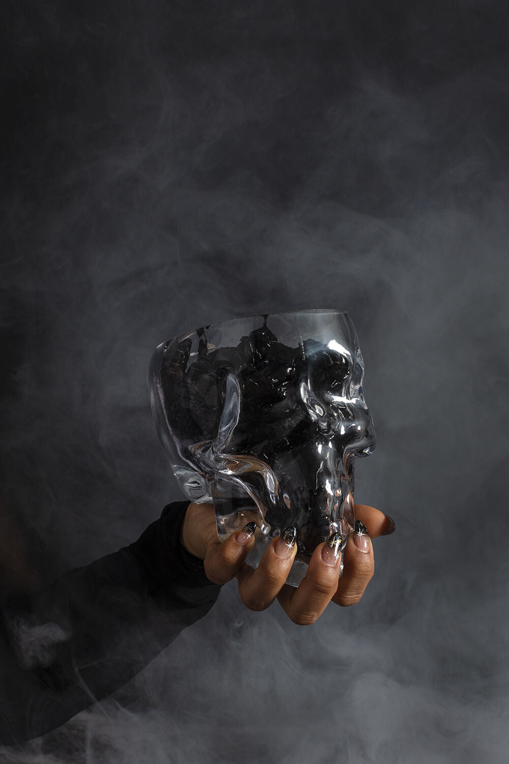 A hand with long, decorated nails holds the Accent Decor Reaper Pot, a transparent skull-shaped terrarium filled with dark liquid against a smoky backdrop—perfect for adding witchy decor vibes.