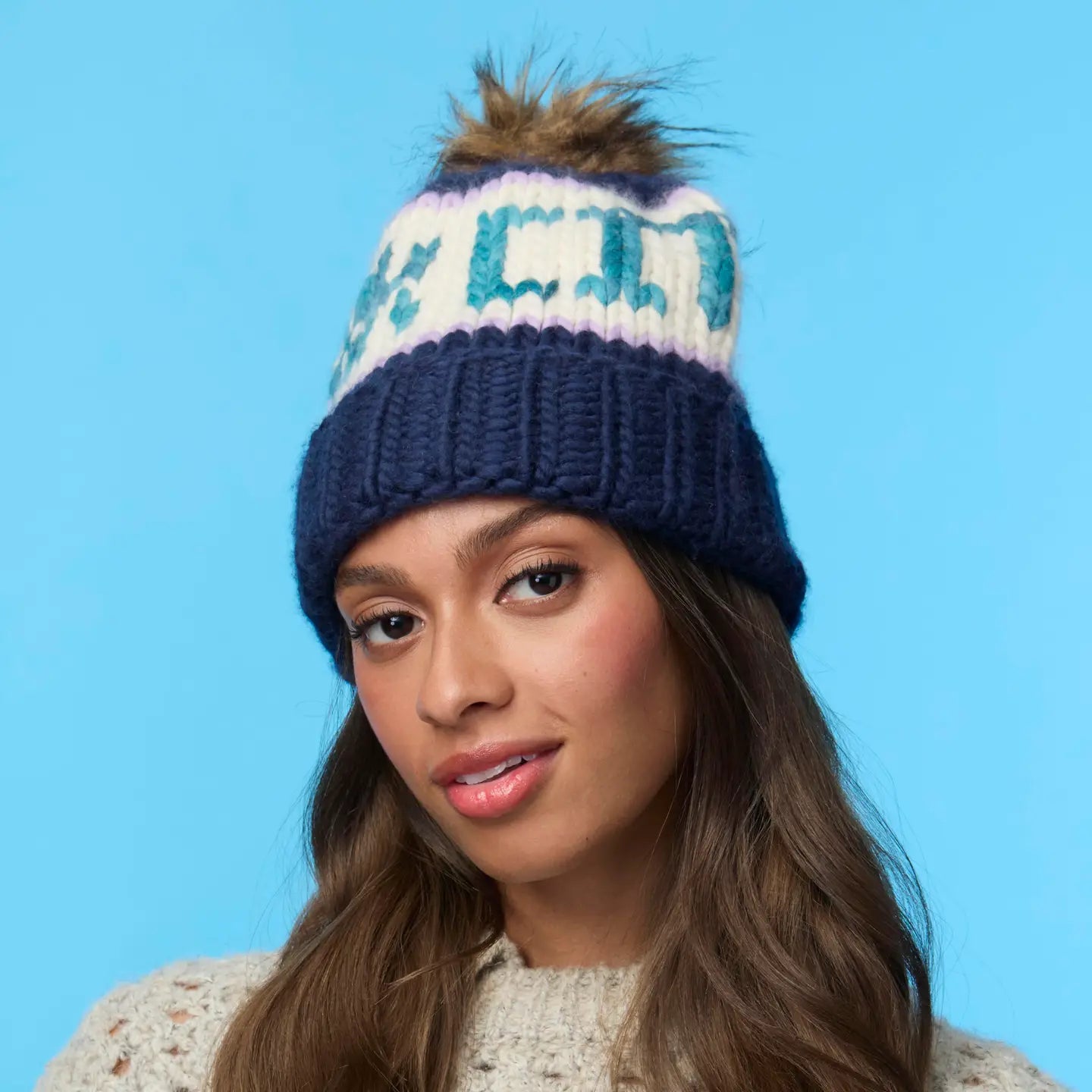 A woman with long brown hair models the Faire Knit Ski Hat, a luxury blue and white beanie with a faux fur pom pom, paired with a cream sweater against a light blue background.