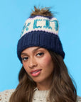 A woman with long brown hair models the Faire Knit Ski Hat, a luxury blue and white beanie with a faux fur pom pom, paired with a cream sweater against a light blue background.