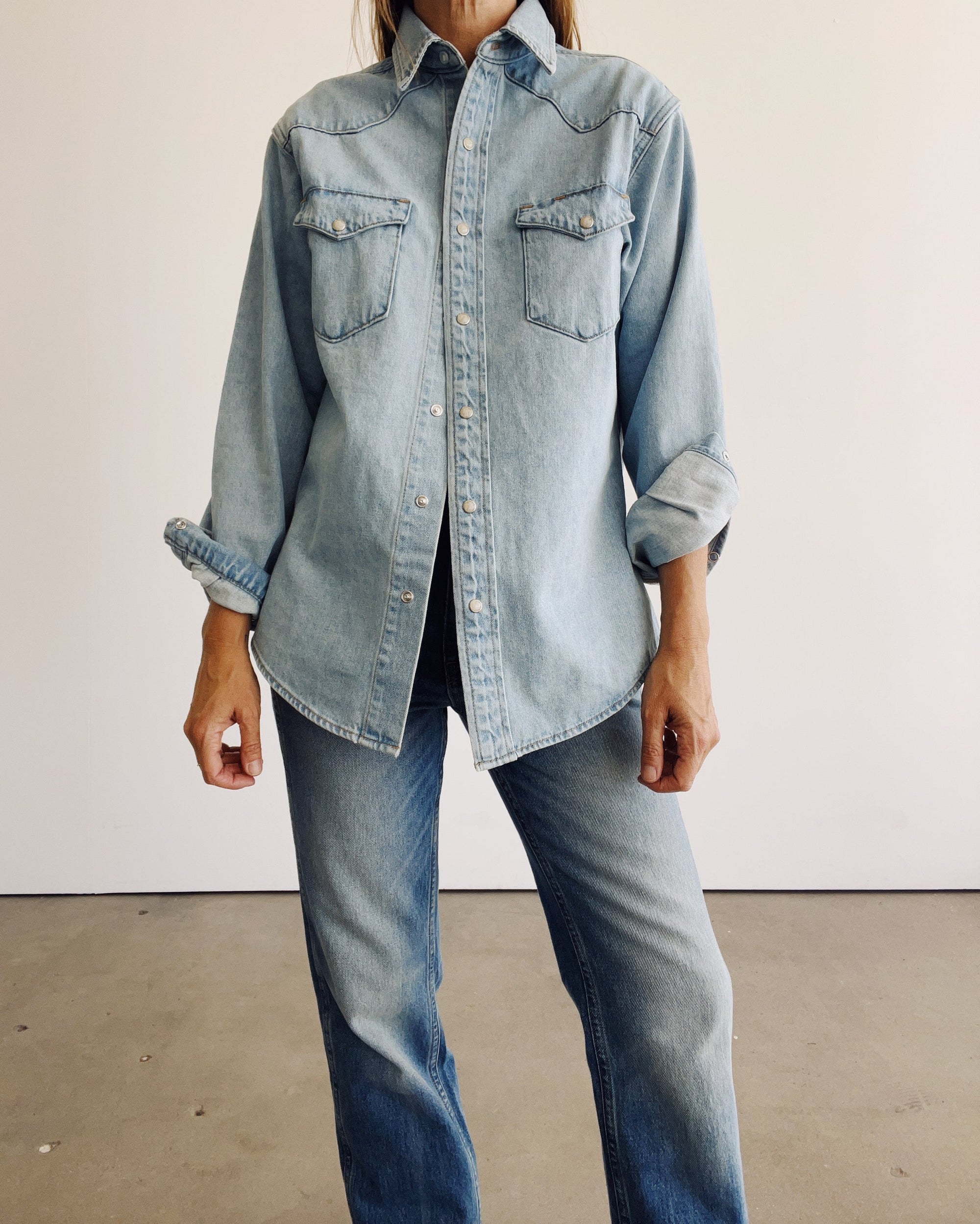 Someone stands indoors against a white wall and concrete floor, wearing The Work Shirt by Hey Gang—a boxy fit light blue denim shirt with rolled-up sleeves—paired with blue jeans.