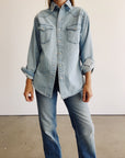 Someone stands indoors against a white wall and concrete floor, wearing The Work Shirt by Hey Gang—a boxy fit light blue denim shirt with rolled-up sleeves—paired with blue jeans.