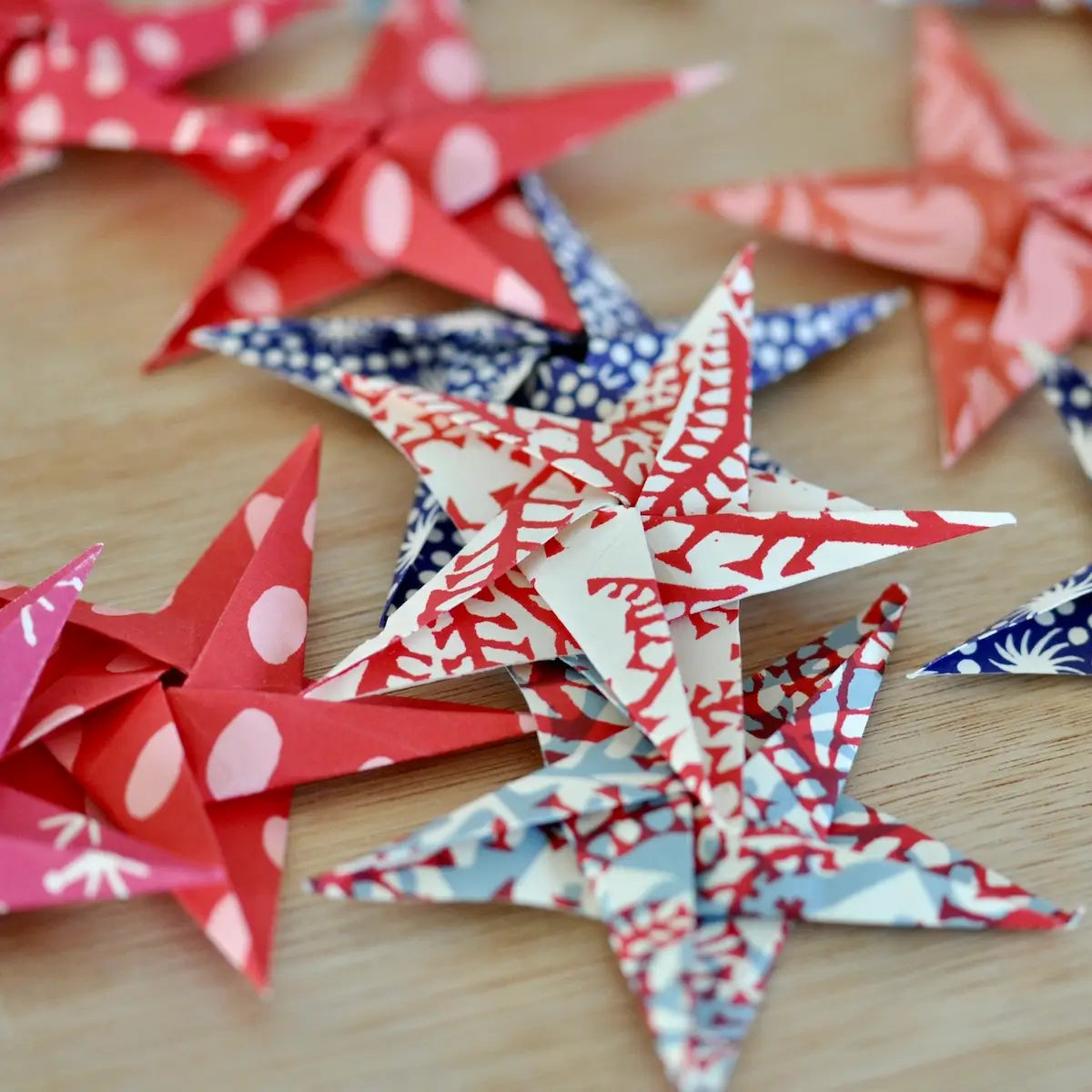 Faire’s Origami Star Garland features red, blue, and white patterned stars made from quality origami paper, artfully displayed on a light wooden surface.