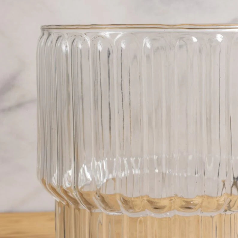 The Shae Champagne Bucket by Be Home features clear, ribbed glass with vertical grooves. Shown on a light wood surface against a neutral background—ideal for luxury entertaining.