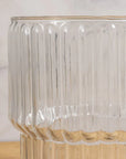 The Shae Champagne Bucket by Be Home features clear, ribbed glass with vertical grooves. Shown on a light wood surface against a neutral background—ideal for luxury entertaining.