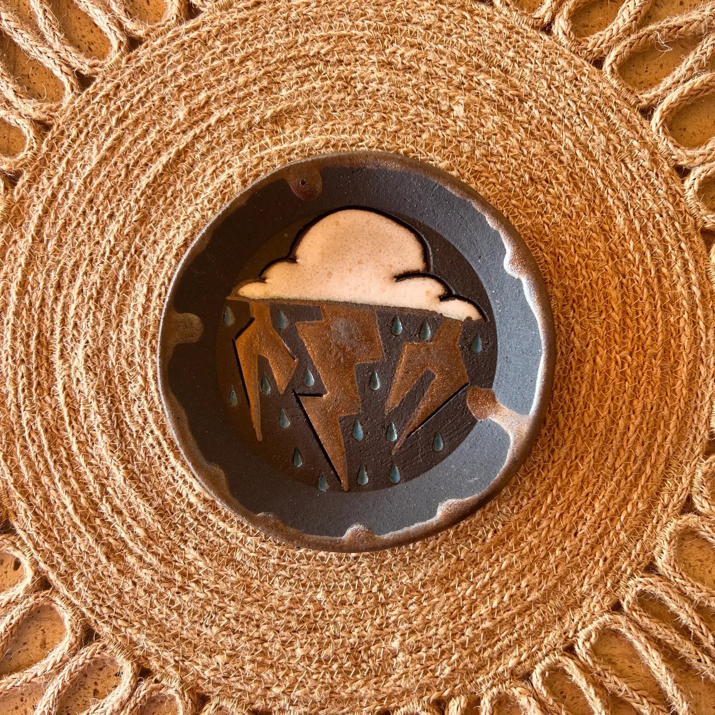 The Monsoon Trinket Dish by Faire features a ceramic plate with a stylized cloud, metallic lightning bolt, and raindrops on a sunburst-patterned placemat—perfect for adding unique flair to your decor.