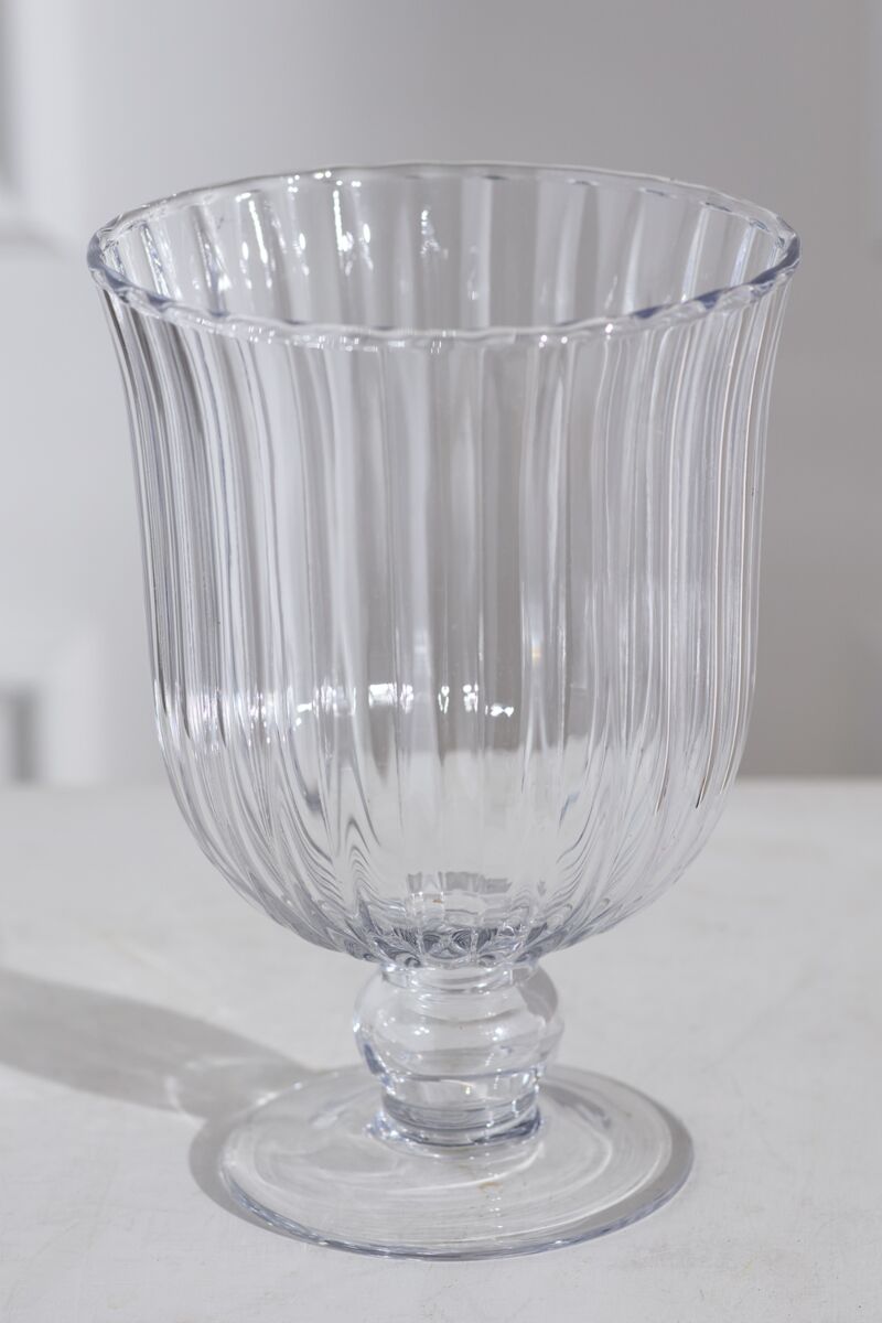 The Oster Vase M by Accent Decor, a clear glass vase with ribbed detailing, a wide flared rim, and a short pedestal base, is displayed on a white surface against a light background.