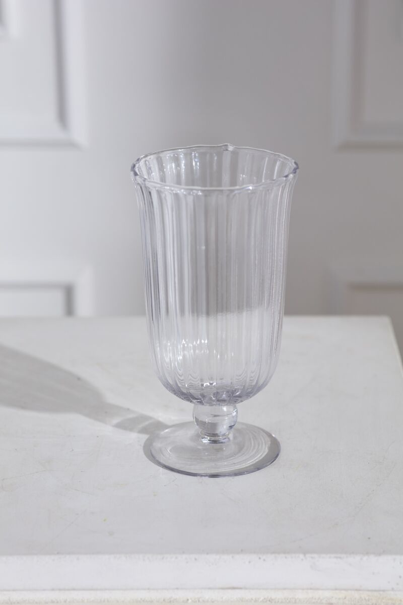 The Oster Vase S by Accent Decor—a clear, fluted glass vase—stands on a white surface, casting a faint shadow. Its elegant, handmade-inspired lines are highlighted against softly lit, white paneled walls in the background.