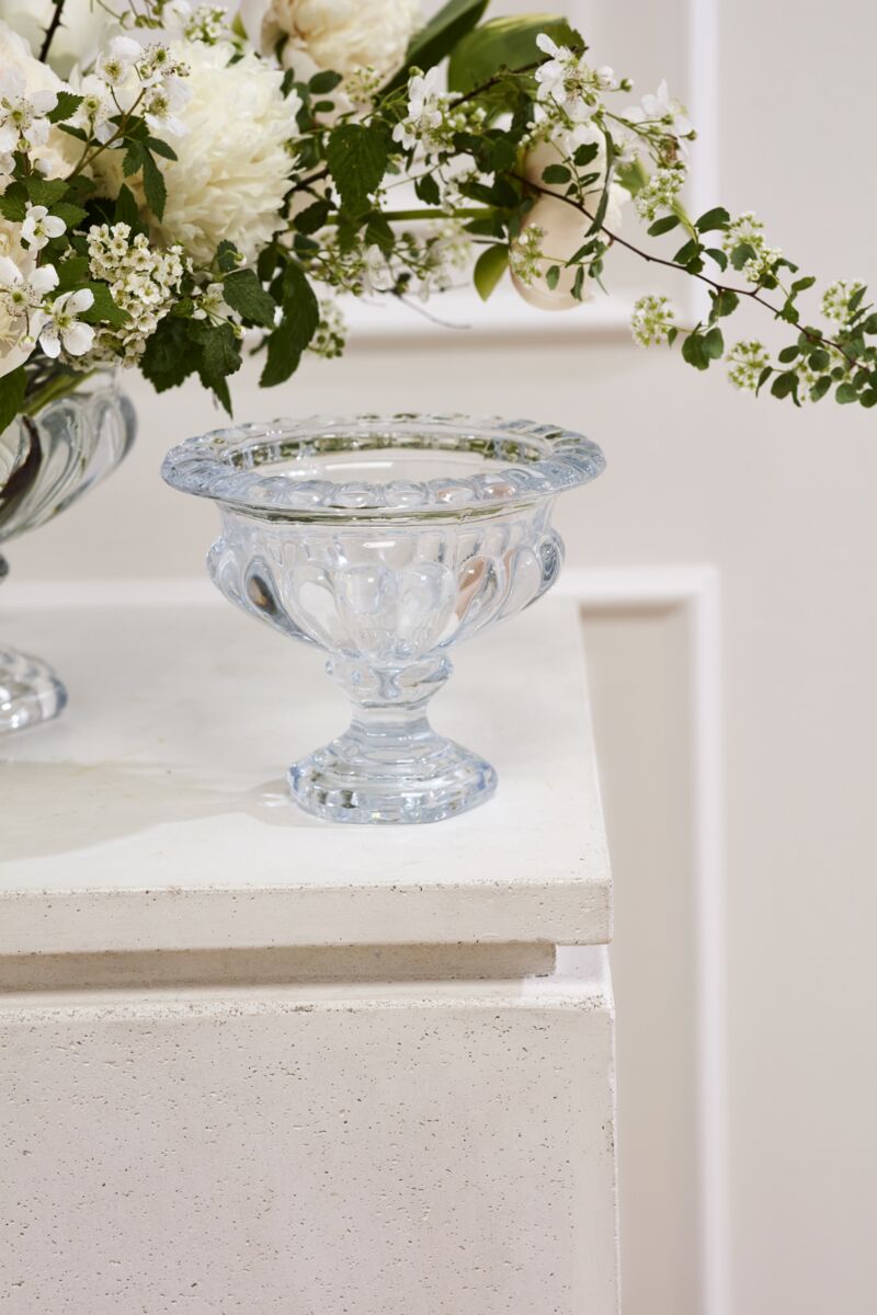The Hathaway Compote S by Accent Decor, a clear vintage glass piece with a wide rim and decorative base, sits on a white textured surface beside a floral arrangement of white flowers and green leaves.