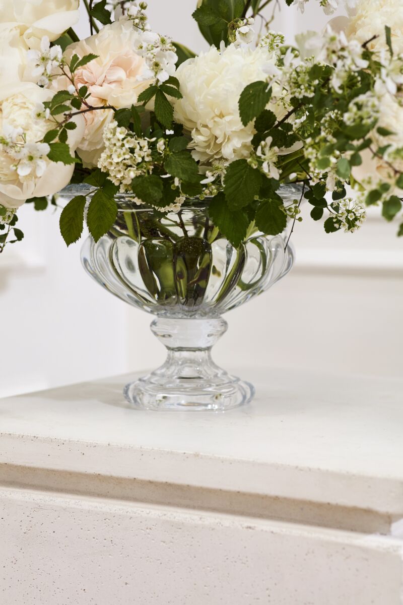 The Hathaway Compote M by Accent Decor holds a bouquet of white and pale peach flowers with green leaves, displayed on a white surface against a light background.