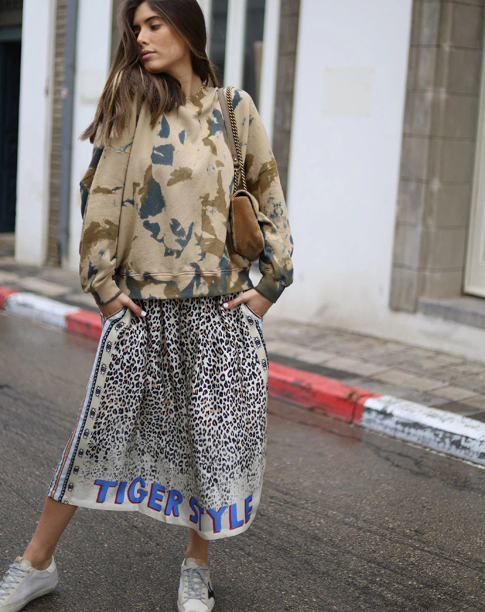 A woman stands on a wet street in a beige and blue camo sweatshirt, Me369's Hailey Printed Skirt with "TIGER STYLE" at the hem, true to size white sneakers, and a brown shoulder bag. She looks to the side.