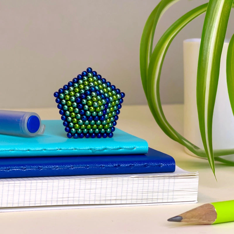 The Faire Speks -512 Gradiante Soothe, a set of blue and green magnet balls, sits on blue notebooks beside a pen, plant, and yellow pencil, adding visual appeal and stress relief to your desk.