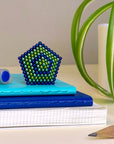 The Faire Speks -512 Gradiante Soothe, a set of blue and green magnet balls, sits on blue notebooks beside a pen, plant, and yellow pencil, adding visual appeal and stress relief to your desk.