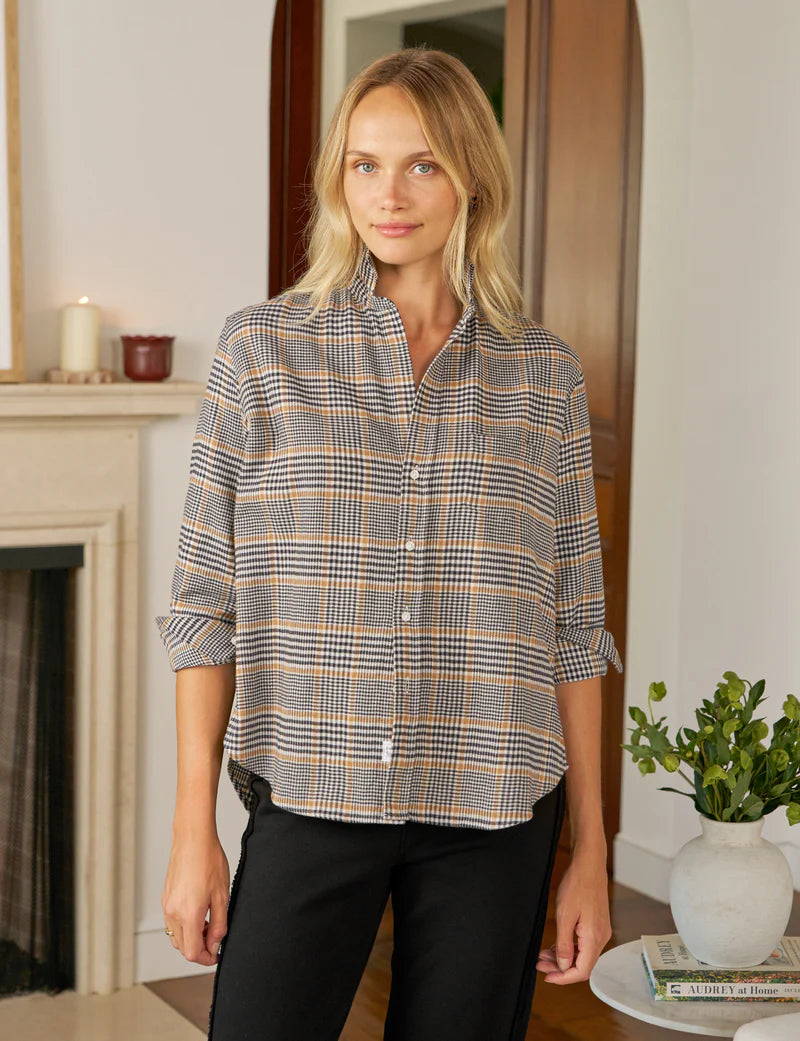 A blonde woman wears the Frank & Eileen Eileen Button Up Shirt F25 with bust-flattering buttons and black pants, standing indoors by a fireplace, near a table with a plant and magazines.